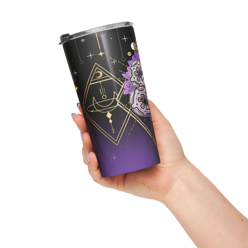 May include: A black and purple travel tumbler with a celestial design. The tumbler features gold geometric shapes, stars, and a crescent moon. A floral design in purple and white is also present. The bottom of the tumbler fades to a deep purple colour.