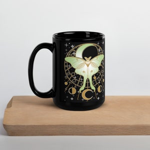 Puede incluir: Taza de cer&aacute;mica negra con un dise&ntilde;o de polilla luna blanca y verde. La polilla est&aacute; delante de una luna creciente con detalles dorados y estrellas. La taza tiene un asa negra.