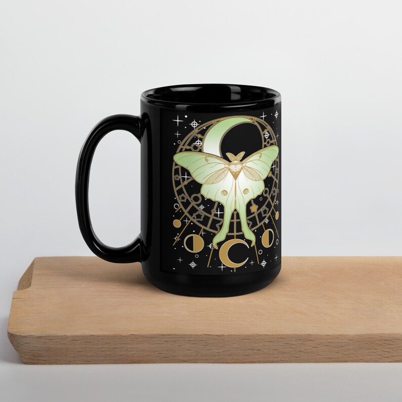 Puede incluir: Taza de cer&aacute;mica negra con un dise&ntilde;o de polilla luna blanca y verde. La polilla est&aacute; delante de una luna creciente con detalles dorados y estrellas. La taza tiene un asa negra.