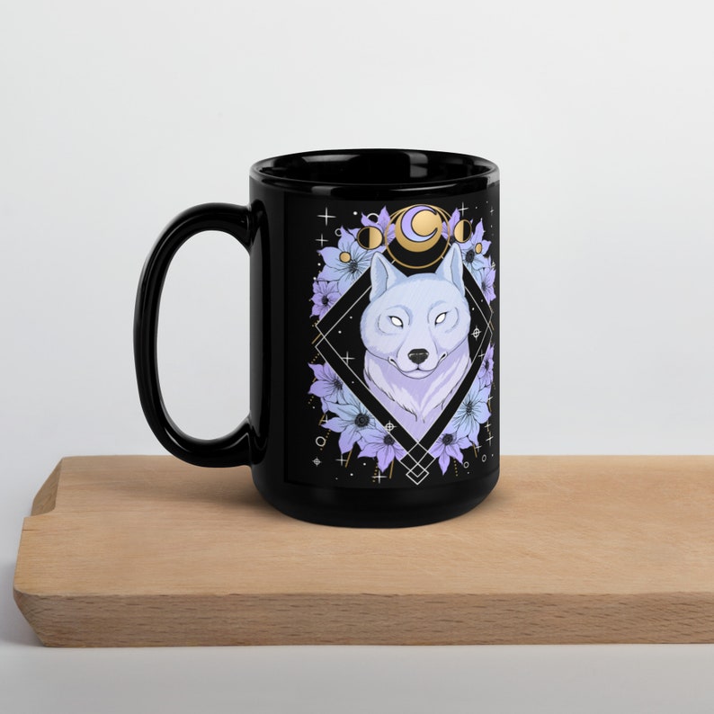 May include: Black ceramic mug featuring a white wolf illustration within a diamond frame, surrounded by purple flowers and celestial elements. The design includes phases of the moon. The mug has a large handle and is displayed on a wooden surface.