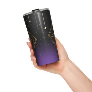 May include: A black and purple travel tumbler with a celestial design. The tumbler has gold accents, stars, and geometric shapes. The bottom of the tumbler fades to a deep purple. The lid is black.