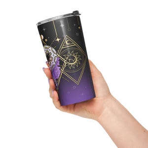 May include: A black and purple travel tumbler with a celestial design. The tumbler features gold geometric shapes, stars, and a sun and moon motif. A floral design in white and purple is also visible.