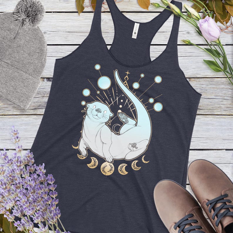 May include: A navy blue tank top with a white and blue illustration of an otter swimming in a circle of moons. The otter is surrounded by a celestial design with stars and dots.