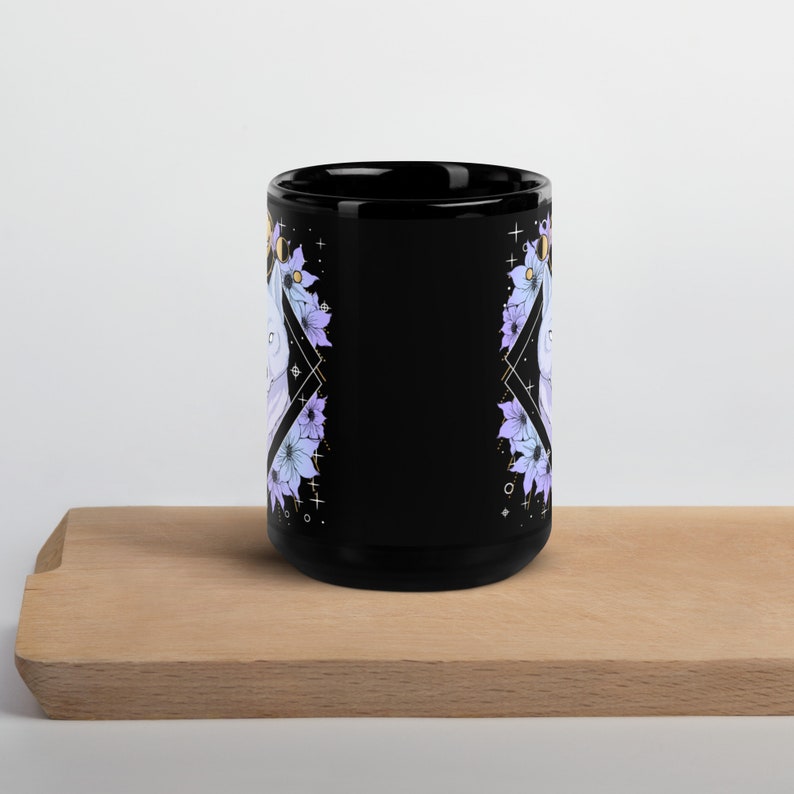 May include: Black ceramic mug featuring a floral design with purple and blue flowers, gold accents, and a geometric pattern. The mug is on a wooden surface.