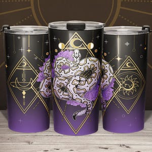 May include: Three black travel mugs with a purple gradient base and gold geometric designs. Each mug features a white and purple snake design, with a crescent moon and sun symbol. The mugs have a black lid and are decorated with stars.