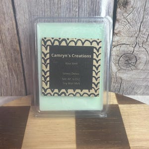 Pode incluir: Um derretimento de cera verde claro em um recipiente de plástico transparente. O rótulo diz "Camryn's Creations" e "Stress Detox". O peso líquido é de 170 gramas. O derretimento de cera é à base de soja.