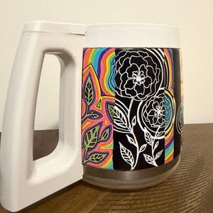 Könnte beinhalten: Ein weißer Kunststoffbecher mit einem umlaufenden Design, das schwarze Felder mit weißen Linienzeichnungen von Blumen und Blättern sowie abwechselnde, leuchtend bunte Regenbogenabschnitte mit grünem Blattwerk und abstrakten Formen zeigt.