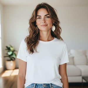 May include: A woman wearing a white crew neck t-shirt and blue jeans. She has long, wavy brown hair and is wearing leaf-shaped earrings. The background is a neutral-toned room with a plant and a white couch.