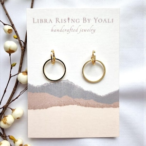 May include: A pair of gold-colored hoop earrings with a small clasp at the top. The earrings are displayed on a white card with the text "LIBRA RISING BY YOALI handcrafted jewelry" and a watercolor design in shades of gray, brown, and pink.