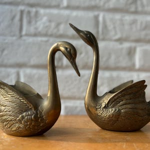 May include: Two brass swan figurines with detailed feather textures on their wings and bodies. One swan is positioned with its head bowed downward, while the other swan has its head held high, both featuring smooth, polished metallic surfaces.