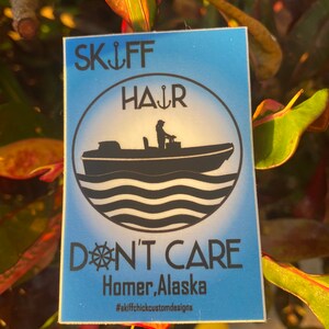 May include: A blue and white sticker with the text "Skiff Hair Don't Care Homer, Alaska #skiffchickcustomdesigns". The sticker features a silhouette of a person in a boat with waves behind it.