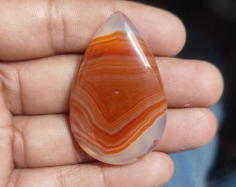 May include: A teardrop-shaped polished gemstone with bands of orange and white. The stone is held in a hand, showcasing its smooth, glossy surface and natural color variations. The gemstone's pattern creates a striking visual effect.