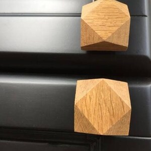 以下が含まれることがあります： ダイヤモンド型の幾何学模様が施された木製のキャビネットノブ2個。ノブは薄い茶色の木でできており、黒い表面に取り付けられています。