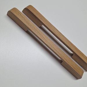 Peut inclure: Deux poignées de meubles en bois avec une finition brun clair. Les poignées ont une forme rectangulaire avec un dessus incurvé et une zone encastrée pour la prise. Elles mesurent environ 25 cm de long.