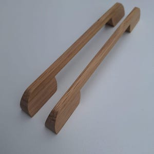 Op de afbeelding: Twee houten kastgrepen met een afgeronde rechthoekige vorm en een lichte bocht aan de bovenkant. De grepen zijn gemaakt van lichtbruin hout.