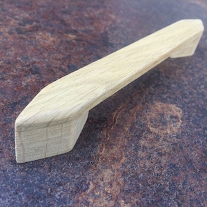 May include: A light brown wooden cabinet pull with a pointed, angled design. The pull is made of natural wood and has a smooth, polished finish.