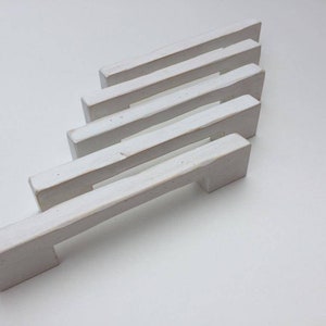 Peut inclure: Cinq poignées de tiroir en céramique blanche de forme rectangulaire avec une légère courbe. Les poignées sont disposées en quinconce, la plus grande en bas et la plus petite en haut.