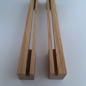 Puede incluir: Dos soportes de teléfono de madera clara. Cada soporte tiene una base rectangular con una ranura y un soporte vertical con una ranura central. Los soportes están diseñados para sostener un teléfono en posición vertical.