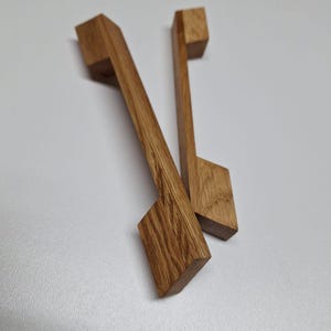 Könnte beinhalten: Zwei rechteckige, leicht angewinkelte Holzknaufgriffe. Die Griffe sind aus hellbraunem Holz mit natürlicher Maserung.