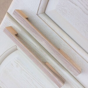 May include: Two wooden cabinet pulls with a light wood grain finish. The pulls are rectangular with rounded edges and a slight curve in the centre.
