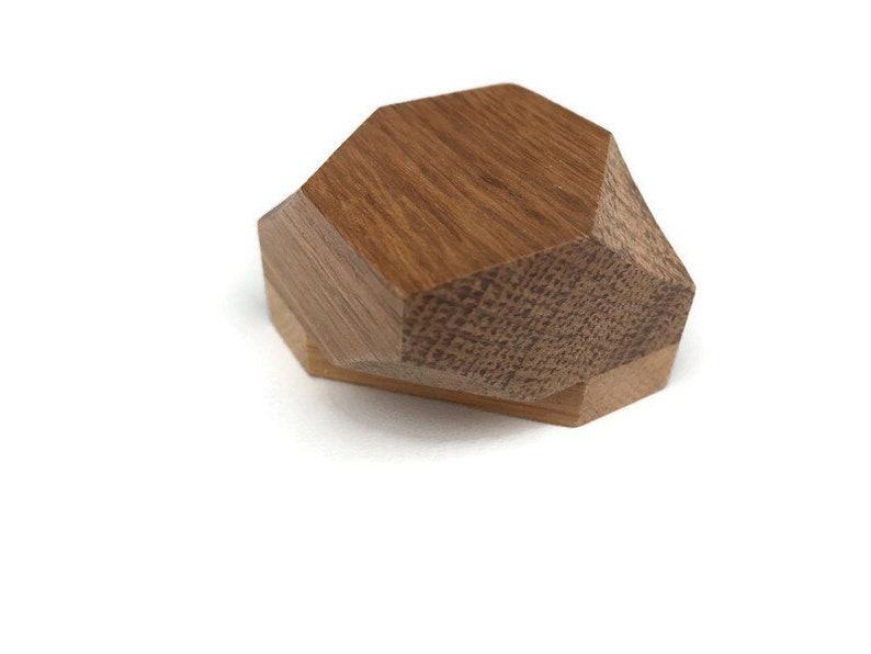 Puede incluir: Una caja geom&eacute;trica de madera con forma hexagonal. La caja est&aacute; hecha de dos piezas de madera, una marr&oacute;n claro y otra marr&oacute;n oscuro. La caja est&aacute; cerrada y tumbada de lado.