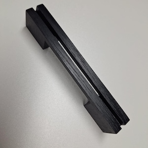 May include: A black cabinet handle with a linear, rectangular design featuring a distinct vertical groove running along the center of the top surface. The handle has two mounting points at each end and a visible wood grain texture throughout.