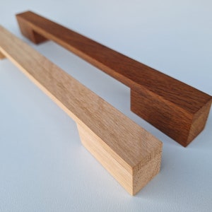 Op de afbeelding: Twee houten kastgrepen, een van naturel hout en een in een donkerbruine kleur gebeitst. Beide grepen hebben een rechthoekige vorm met een uitsparing in het midden.