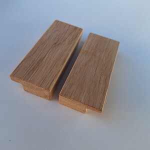 May include: Two wooden drawer pulls with a natural finish. The pulls are rectangular with a slight curve at the top and a small notch at the bottom.