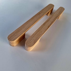 Könnte beinhalten: Zwei Holzknaufgriffe mit natürlicher Oberfläche. Die Griffe sind rechteckig mit abgerundeten Kanten und einer leichten Wölbung in der Mitte.