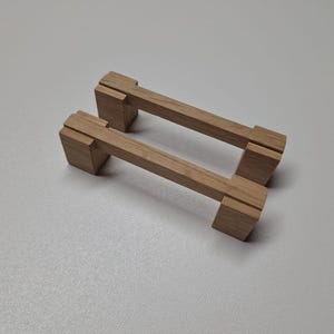 Könnte beinhalten: Zwei Holzschrankgriffe. Jeder Griff hat einen rechteckigen Griff und zwei quadratische Endstücke. Das Holz hat eine hellbraune Farbe und eine sichtbare Maserung. Die Griffe sind für eine einfache Installation an Schubladen oder Schränken konzipiert.