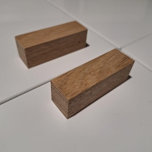 May include: Two wooden blocks, both rectangular in shape, with a light brown color and a wood grain texture. The blocks are lying flat on a white surface.
