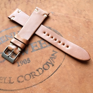 May include: A light brown leather watch strap with cream-colored stitching and a silver buckle. The strap is made of a smooth, high-quality leather and is designed to fit a standard watch. The strap is shown on a brown leather surface.