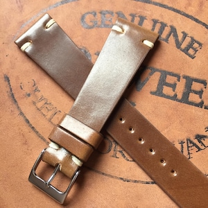 Peut inclure: Un bracelet de montre en cuir marron lisse avec des coutures de couleur crème près des cornes et une boucle en métal argenté poli. Le bracelet se compose de deux parties, l'une avec une série de trous perforés et l'autre avec la boucle.
