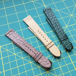 May include: Three leather watch straps in different colors: brown, beige, and green. The straps have a textured alligator pattern and are laid out on a green grid.