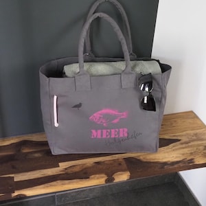 Könnte beinhalten: Graue Canvas-Tragetasche mit einem rosa Fisch-Motiv und dem Wort "MEER" in Rosa. Die Tasche hat zwei Griffe, eine Seitentasche mit rosa Reißverschluss und eine Sonnenbrille. Ein gefaltetes Handtuch befindet sich im Inneren.