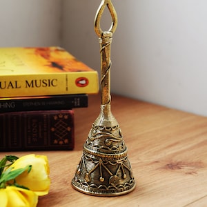 Pu&ograve; includere: Una campana di ottone dettagliata con un manico ad anello aperto. La campana presenta un'intricata filigrana e motivi geometrici. La campana &egrave; esposta su una superficie di legno con libri e rose gialle sullo sfondo.