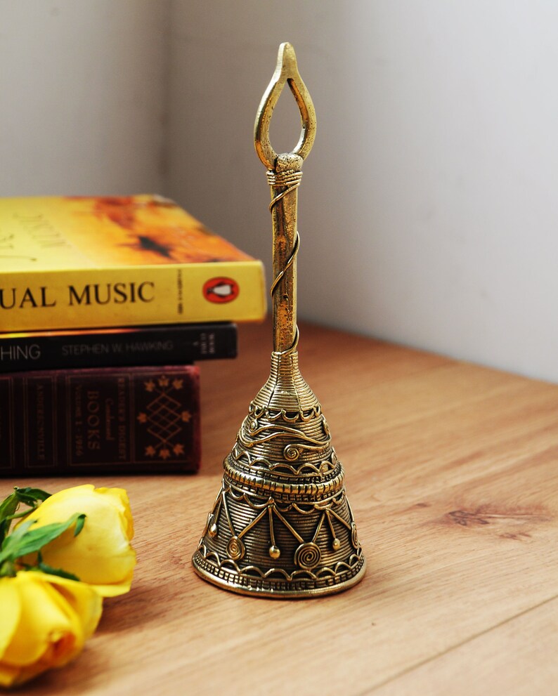Pu&ograve; includere: Una campana di ottone dettagliata con un manico ad anello aperto. La campana presenta un'intricata filigrana e motivi geometrici. La campana &egrave; esposta su una superficie di legno con libri e rose gialle sullo sfondo.