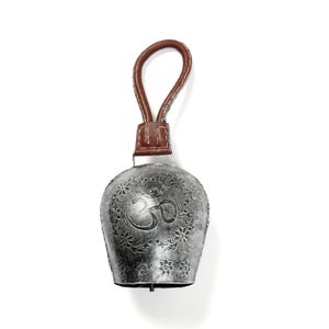 May include: Silver-colored bell with a brown leather handle. The bell features an embossed Om symbol and floral design. The handle is attached to a brown leather band at the top of the bell. The bell is likely made of metal.