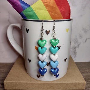 May include: A pair of dangle earrings with a rainbow of heart-shaped beads. The earrings are green, turquoise, white, light blue, and dark blue. The earrings are hanging from a white mug with a rainbow flag on top.