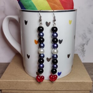 May include: A pair of dangle earrings with a black, white, blue, and purple bead pattern. The earrings have a red heart charm at the bottom and are hanging from a white mug with black hearts.
