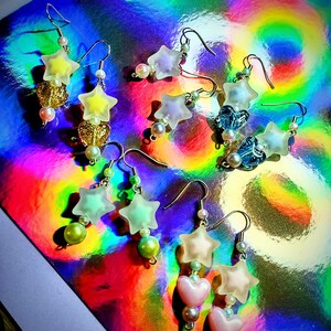 May include: A collection of handmade earrings featuring star and heart-shaped charms. The earrings are in various colors, including yellow, pink, and blue, with pearl and bead accents. The earrings are displayed on a holographic surface.