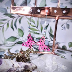 May include: A pair of pink butterfly earrings with silver hooks and beaded accents. The earrings are displayed on a rose gold earring stand against a backdrop of foliage and decorative elements.
