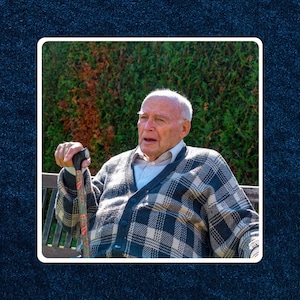May include: An older man with white hair and a plaid sweater sits on a bench outdoors. He is holding a walking stick with a black handle and a colorful design.