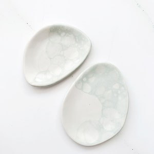 Puede incluir: Dos platos de cerámica blanca con un diseño marmoleado verde claro. Los platos son de forma ovalada y tienen un borde ligeramente irregular.