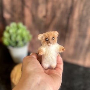 May include: A small, needle-felted bear figurine with a light tan and cream-coloured coat. The bear has two small, dark, round eyes, a tiny nose, and rounded ears. Its arms are extended outwards, and it has a soft, fuzzy texture throughout.