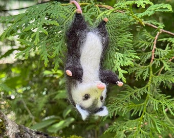 Könnte beinhalten: Eine aus Nadelfilz gefertigte Opossum-Figur, die kopfüber an ihrem rosa Schwanz hängt. Das Opossum hat ein weißes Gesicht mit schwarzen Abzeichen um die Augen, einen weißen Bauch und dunkelgraues Fell an Rücken und Gliedmaßen. Es hat kleine rosa Pfoten.