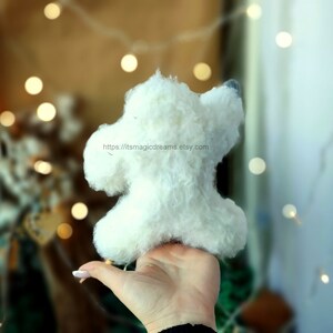 May include: A white fluffy handmade toy dog with a gray nose, held in a hand against a blurred background of lights.