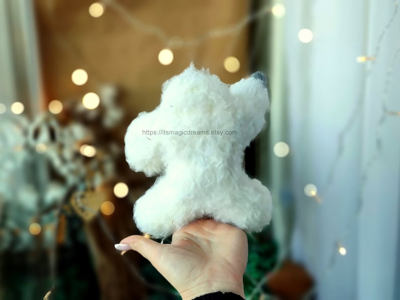 May include: A white fluffy handmade toy dog with a gray nose, held in a hand against a blurred background of lights.