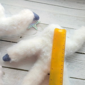 May include: Two white fluffy felt animal figures with blue felt accents and iridescent star embellishments. A yellow ruler is placed next to the figures for size reference.