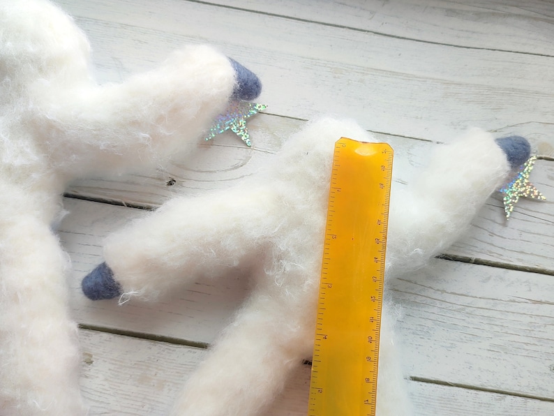 May include: Two white fluffy felt animal figures with blue felt accents and iridescent star embellishments. A yellow ruler is placed next to the figures for size reference.